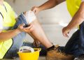 Trabajador con chaleco amarillo sentado en el piso se toma la rodilla lastimada; un compañero lo asiste y hay un casco al lado