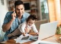 Padre hablando por celular con su beba en brazos frente a una notebook en la mesa de su casa